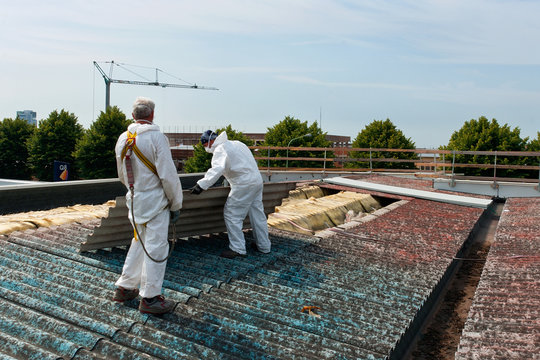 Two workers in white protective suits are removing or working on asbestos roofing materials on a building rooftop, with construction cranes visible in the background.